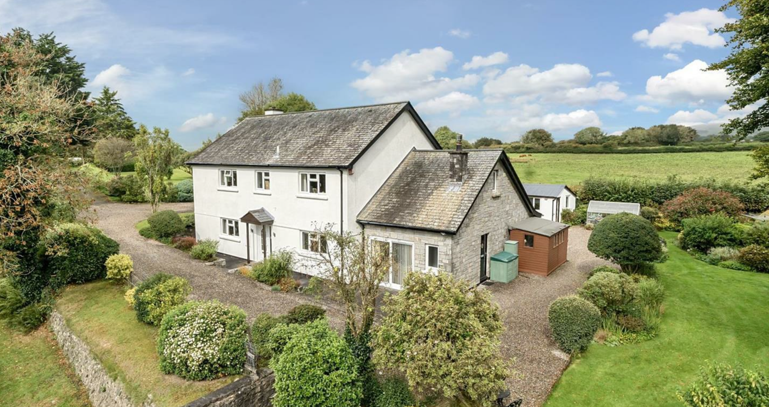A white rendered 1930s detached house with a granite-clad side extension, set in green fields and mature landscaped gardens.