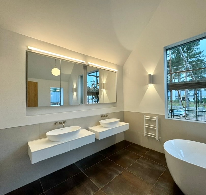 Large bathroom with a vaulted ceiling, freestanding bath and dual sinks mounted on white vanity units. A large window looks out towards trees.