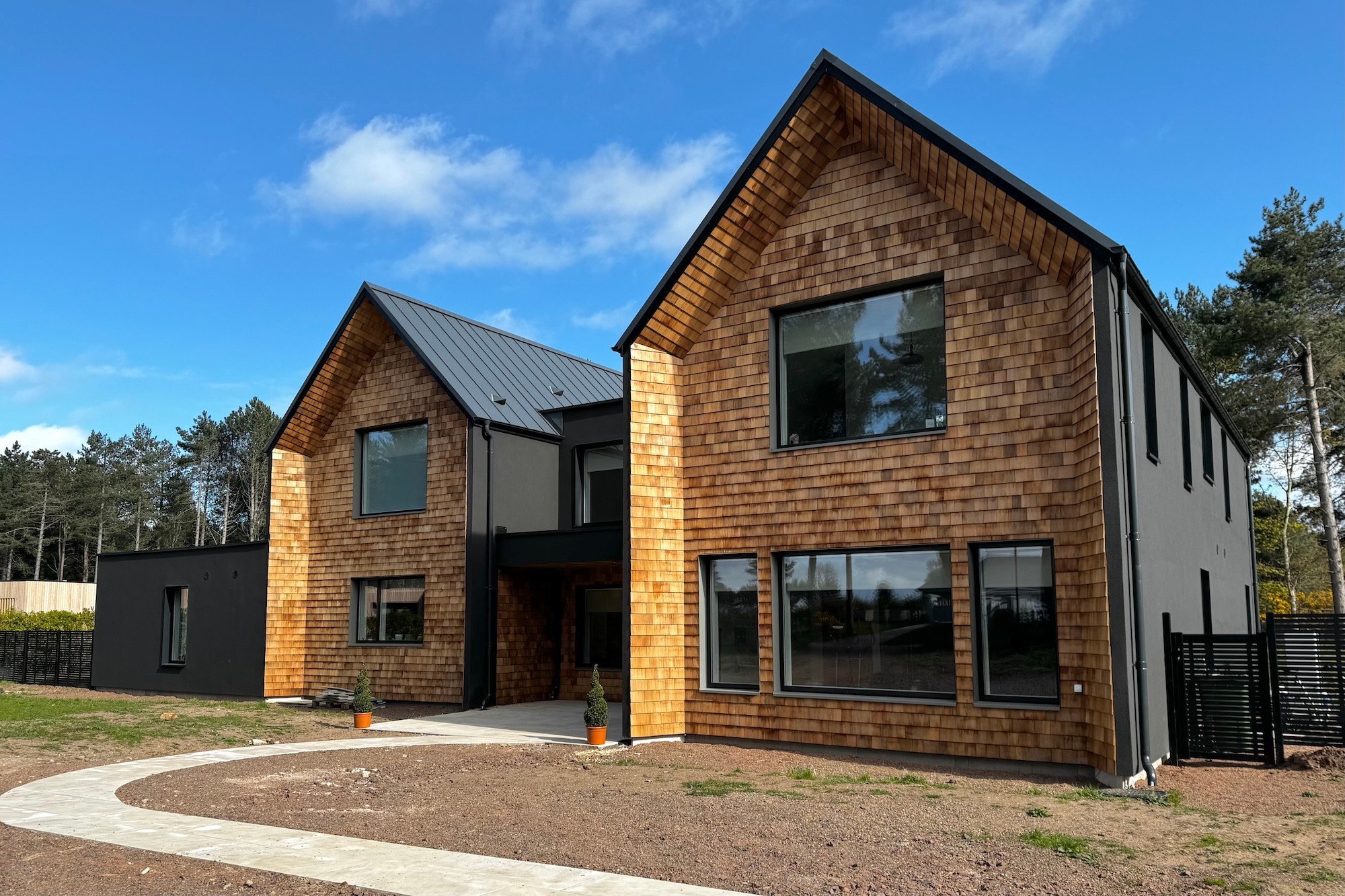 A newly built modern house with two gables, dark zinc roof, black render and cedar shingle gable-ends.