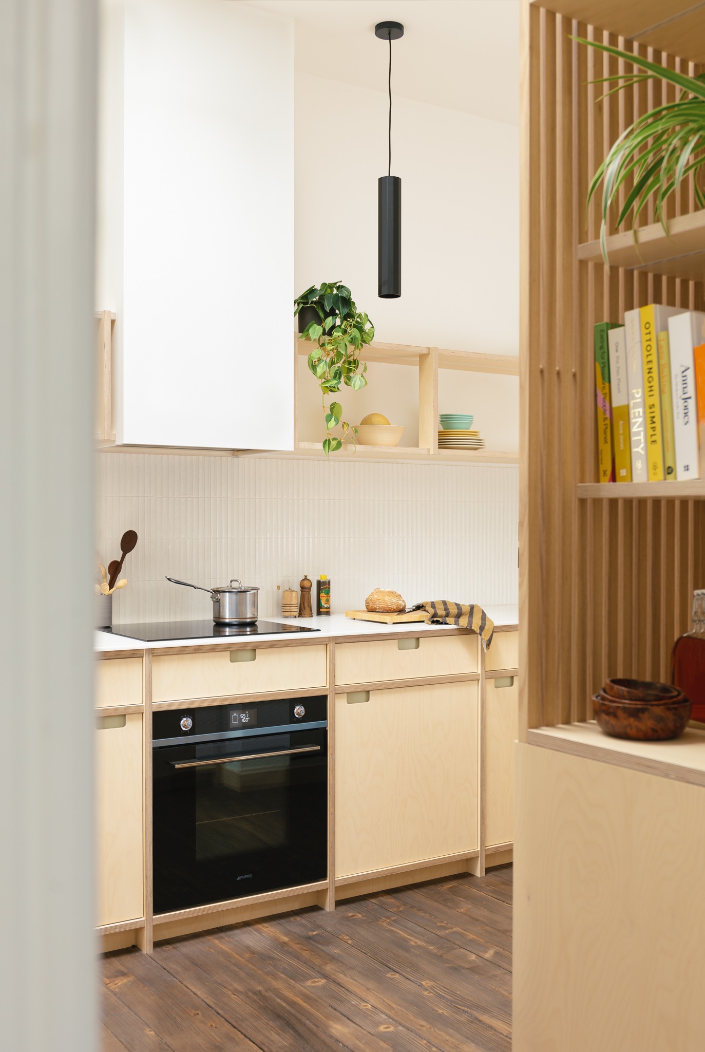 Glimpse of a birch plywood kitchen past shelving unit with slatted ash wood sides, towards an oven and hob with black pendant and open shelves above.